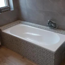 Wykończona łazienka z białą wanną obudowaną mozaiką, szare płytki na ścianach, biała szafka z umywalką i grzejnik żeberkowy.