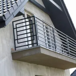 Czarna, metalowa balustrada balkonowa o geometrycznym wzorze, zamontowana na balkonie budynku z szarą elewacją i ciemnym dachem.