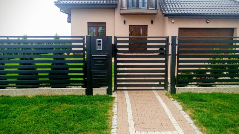 Nowoczesne, poziome ogrodzenie panelowe w kolorze antracytowym z furtką i wideodomofonem, Gryfino. Brukowana ścieżka prowadzi do domu z beżową elewacją i balkonem.