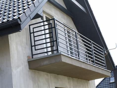 Czarna, metalowa balustrada balkonowa o geometrycznym wzorze, zamontowana na balkonie budynku z szarą elewacją i ciemnym dachem.