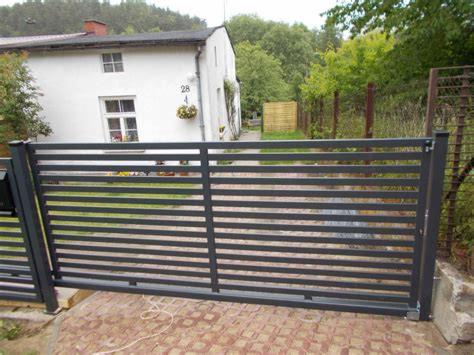 Nowoczesna, pozioma brama wjazdowa z metalowych profili, pomalowana na grafitowo, na tle białego domu z numerem 28 i ogrodu.