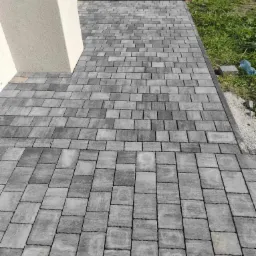 Ułożona szara kostka brukowa tworząca ścieżkę wzdłuż budynku, z widocznymi różnicami w odcieniach poszczególnych kostek i krawężnikiem oddzielającym ścieżkę od trawy.