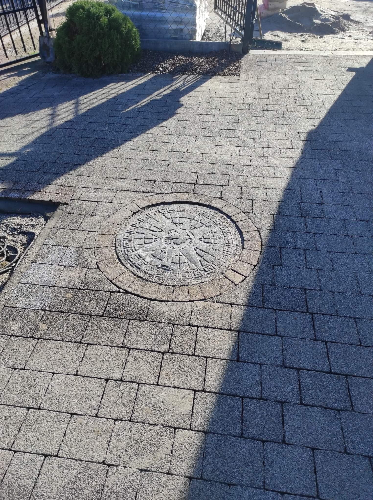 Szara kostka brukowa z dekoracyjnym okrągłym elementem z motywem azteckim pośrodku, oświetlona słońcem i częściowo zacieniona, z widocznym krzewem i elementami ogrodzenia w tle.