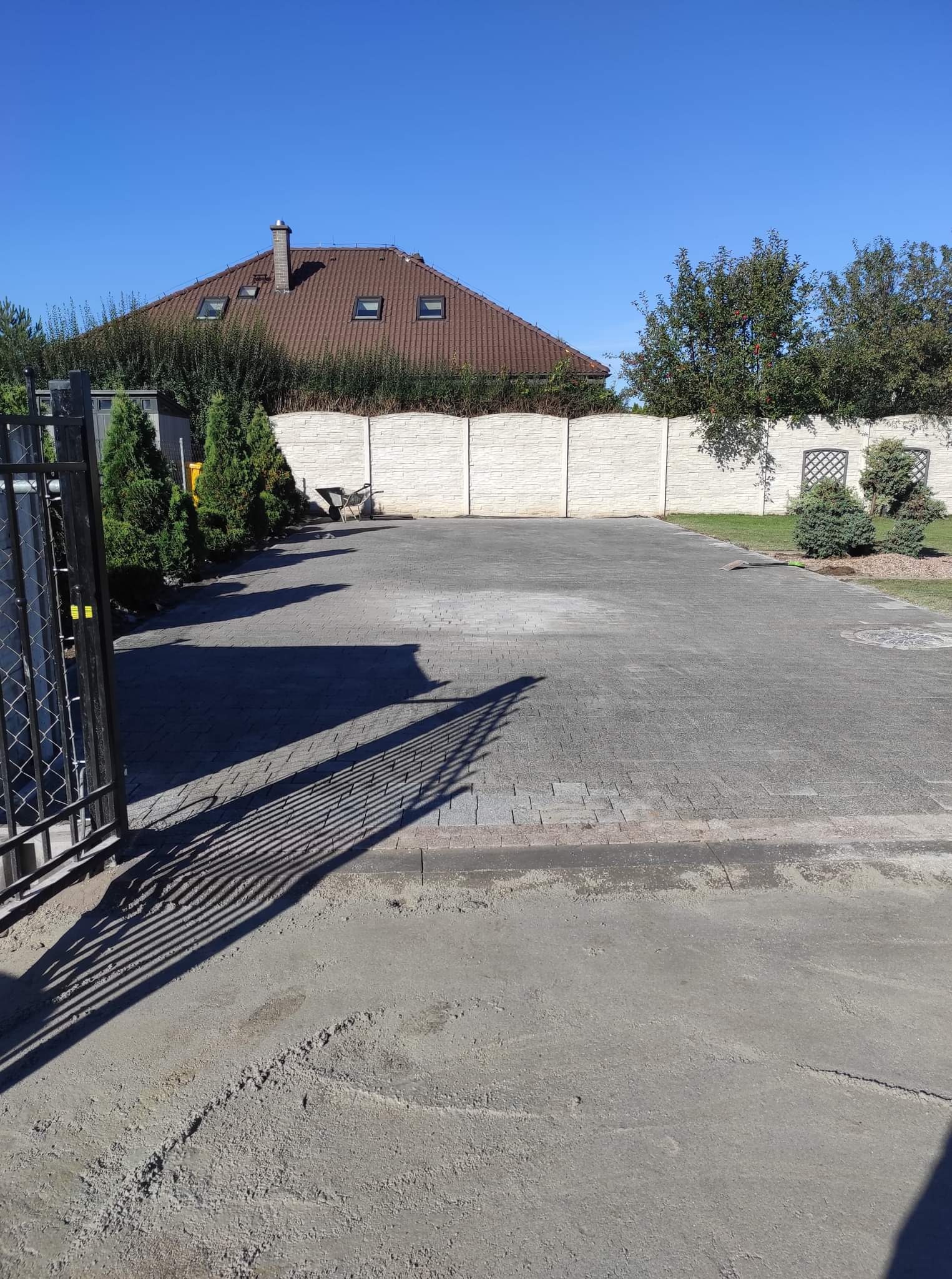Ułożona szara kostka brukowa na podjeździe z widocznymi śladami cementu, ogrodzenie z białych prefabrykatów betonowych, cień metalowej bramy w lewym dolnym rogu.