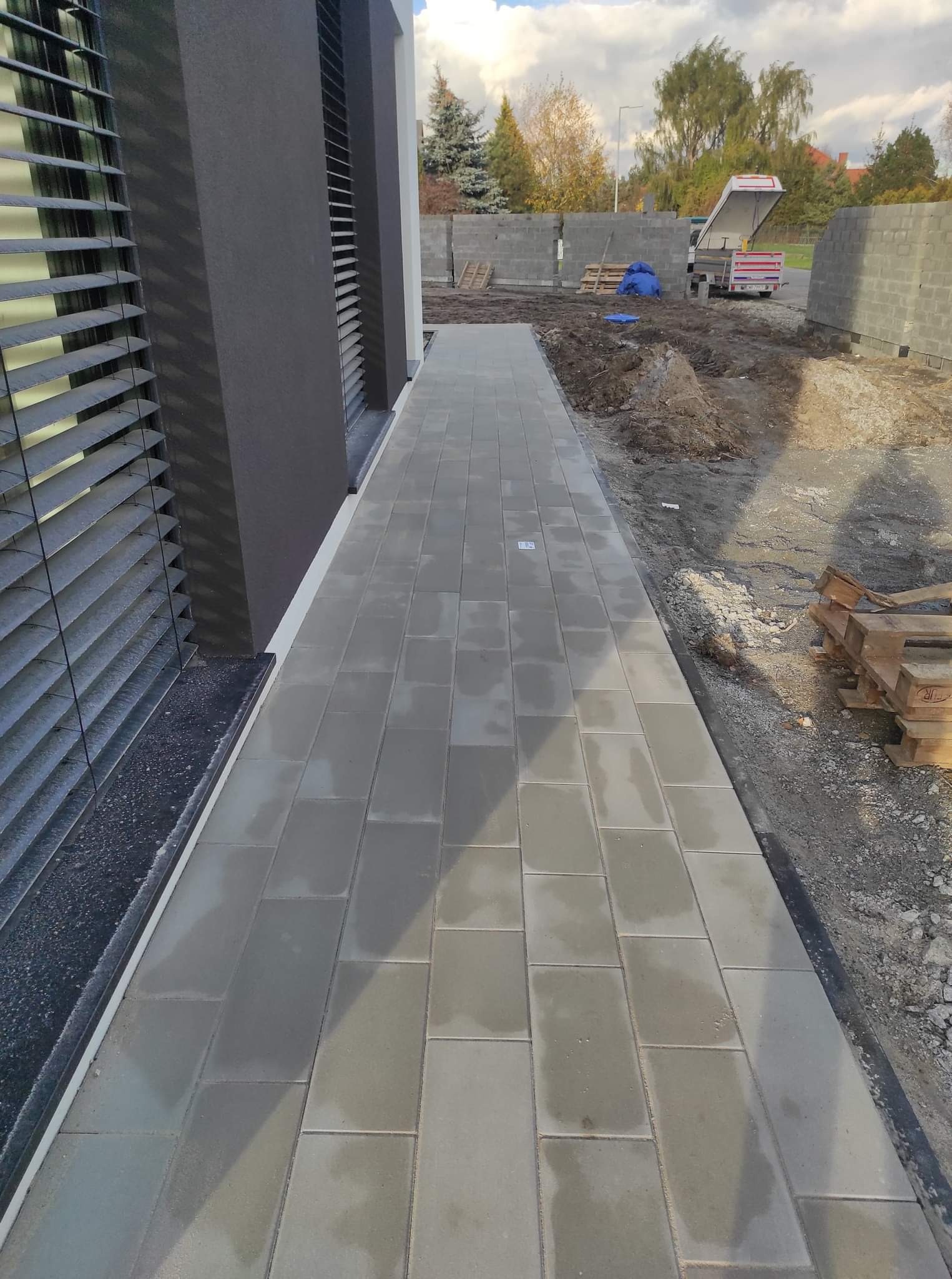 Świeżo ułożona, szara kostka brukowa prowadząca wzdłuż ciemnej elewacji budynku z żaluzjami; w tle plac budowy z bloczkami i zaparkowanym samochodem dostawczym.