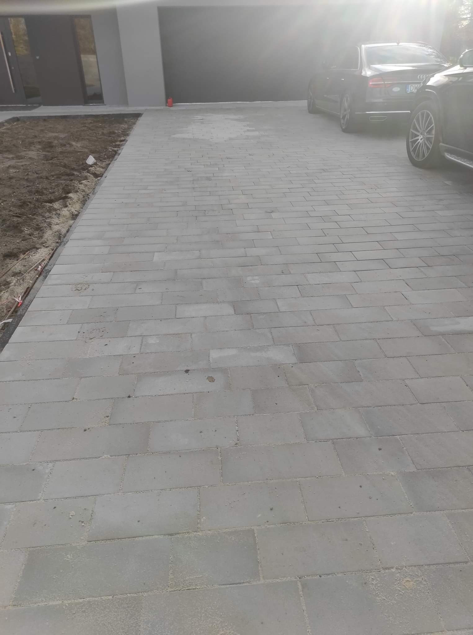 Nowo ułożona, szara kostka brukowa na podjeździe, z zaparkowanymi samochodami w tle i przygotowanym terenem pod trawnik po lewej stronie.