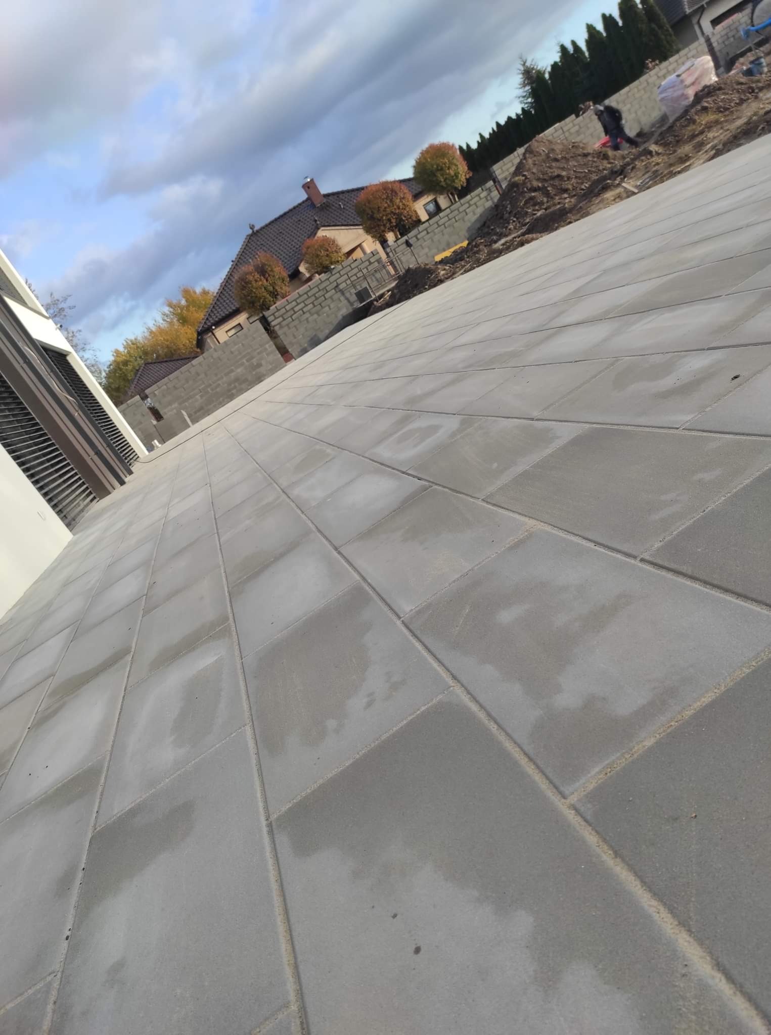 Świeżo ułożona, mokra, szara kostka brukowa na podjeździe domu jednorodzinnego, widok z niskiej perspektywy, w tle budowa ogrodzenia z bloczków betonowych.