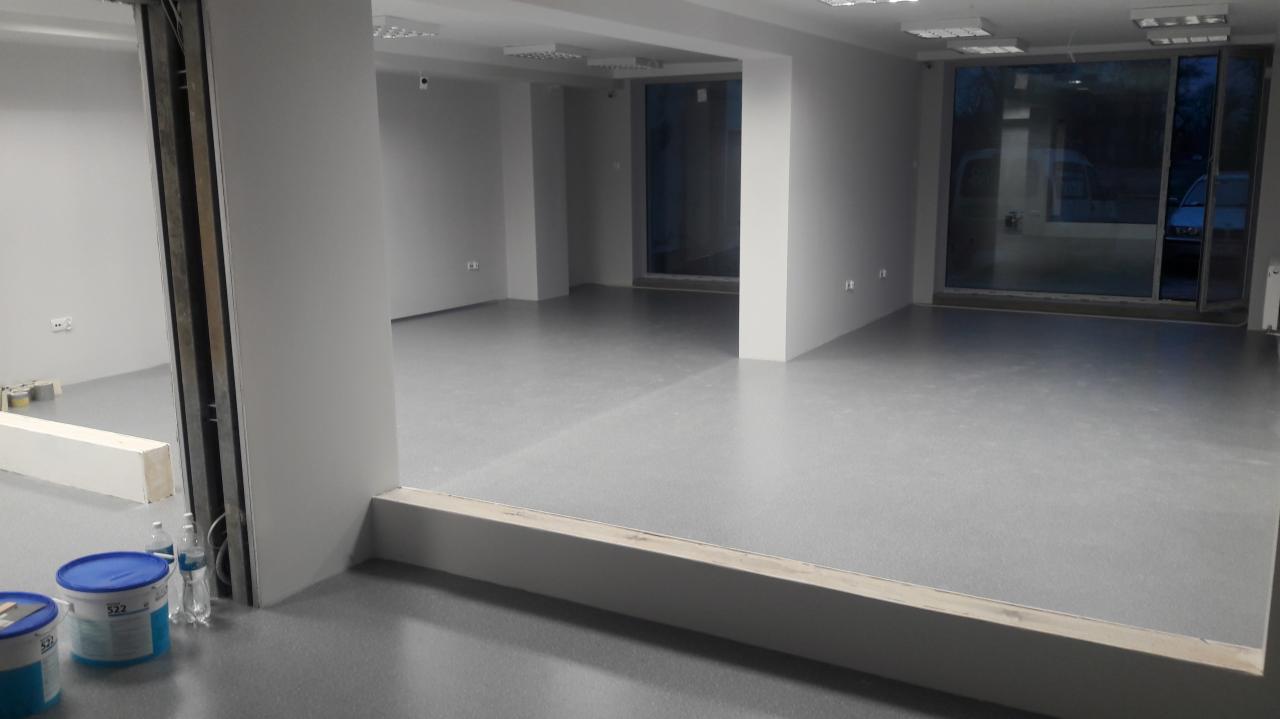 Pomalowane na szaro puste pomieszczenie w trakcie remontu, widoczne puszki z farbą i okno z widokiem na zewnątrz.