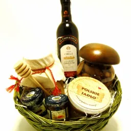 Zielony wiklinowy kosz wypełniony zestawem upominkowym: butelka czerwonego wina El Emperador, słoiczki z dżemem, miód z drewnianym nabierakiem, słoik z orzechami przykryty drewnianym grzybkiem.