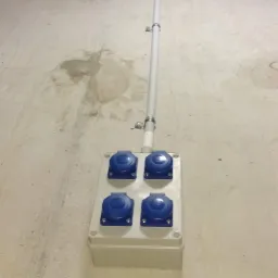 Cztery gniazda elektryczne przemysłowe z niebieskimi pokrywami, zamontowane w szarej skrzynce na betonowej ścianie, z rurą prowadzącą do sufitu.