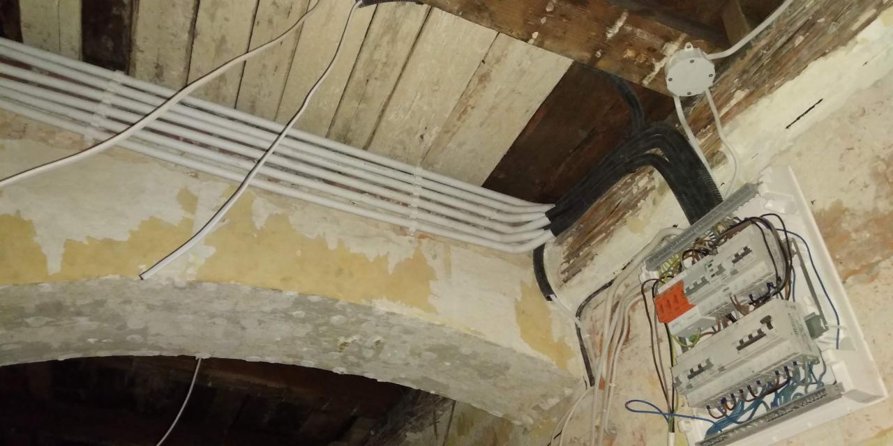 Nowa instalacja elektryczna z białymi rurkami PCV prowadzona po starym, łukowym suficie z drewnianymi belkami i z zamontowaną białą skrzynką rozdzielczą z bezpiecznikami na ścianie z łuszczącą się...