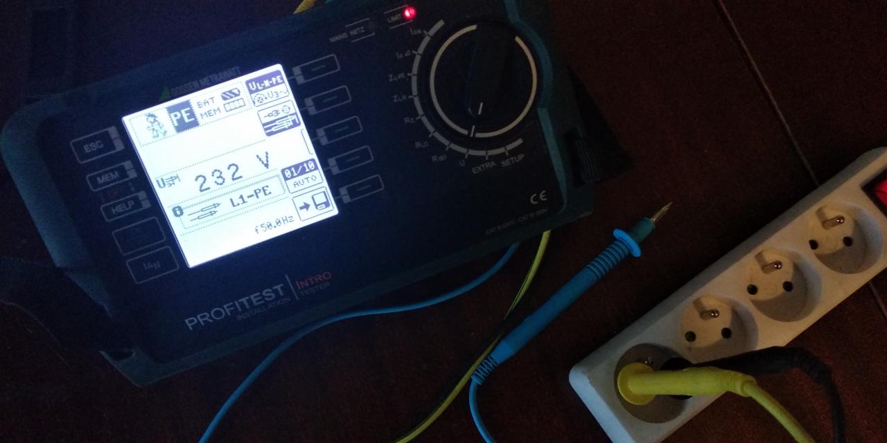 Pomiar napięcia 232V miernikiem PROFITEST na listwie zasilającej z podłączonym przewodem, widoczny wyświetlacz urządzenia i sonda pomiarowa.