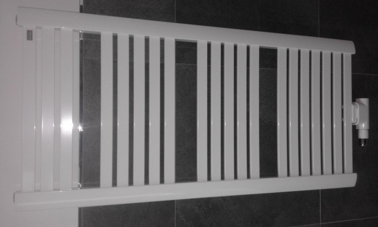 Biały, drabinkowy grzejnik łazienkowy zamontowany na ścianie z ciemnymi, szarymi płytkami, widoczne zawory termostatyczne.
