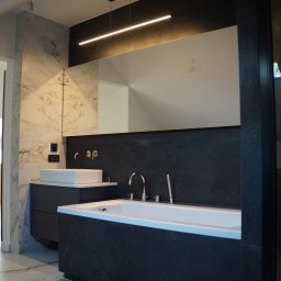 Pillar sp. z o.o. - Nowoczesna łazienka z marmurowymi płytkami na ścianie i podłodze, dużą wanną obudowaną czarnymi płytkami, umywalką nablatową na czarnej szafce i podłużnym oświetleniem LED nad lustrem.