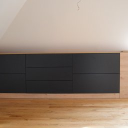 Pillar sp. z o.o. - Czarne, matowe fronty szafy wnękowej pod skosem, z drewnianymi elementami wykończeniowymi i gniazdkami elektrycznymi po prawej stronie, na tle drewnianej podłogi.