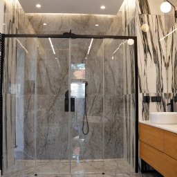 Pillar sp. z o.o. - Nowoczesna łazienka z kabiną prysznicową typu walk-in, wykończona płytkami imitującymi marmur w odcieniach szarości i bieli, z czarną armaturą i drewnianą szafką pod umywalką.