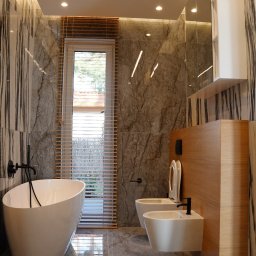 Pillar sp. z o.o. - Nowoczesna łazienka z marmurowymi płytkami, wolnostojącą wanną, podwieszaną toaletą i bidetem, oświetleniem LED i drewnianymi żaluzjami w oknie.