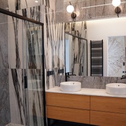 Pillar sp. z o.o. - Nowoczesna łazienka z marmurowymi płytkami, kabiną prysznicową z czarnymi ramami, drewnianą szafką pod umywalki i okrągłymi lampami ściennymi