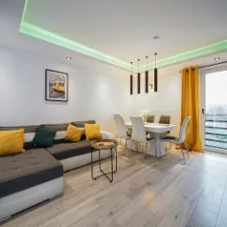 Jasne wnętrze salonu z narożną sofą, stołem jadalnym i oknem balkonowym z żółtymi zasłonami, podświetlenie LED na suficie.