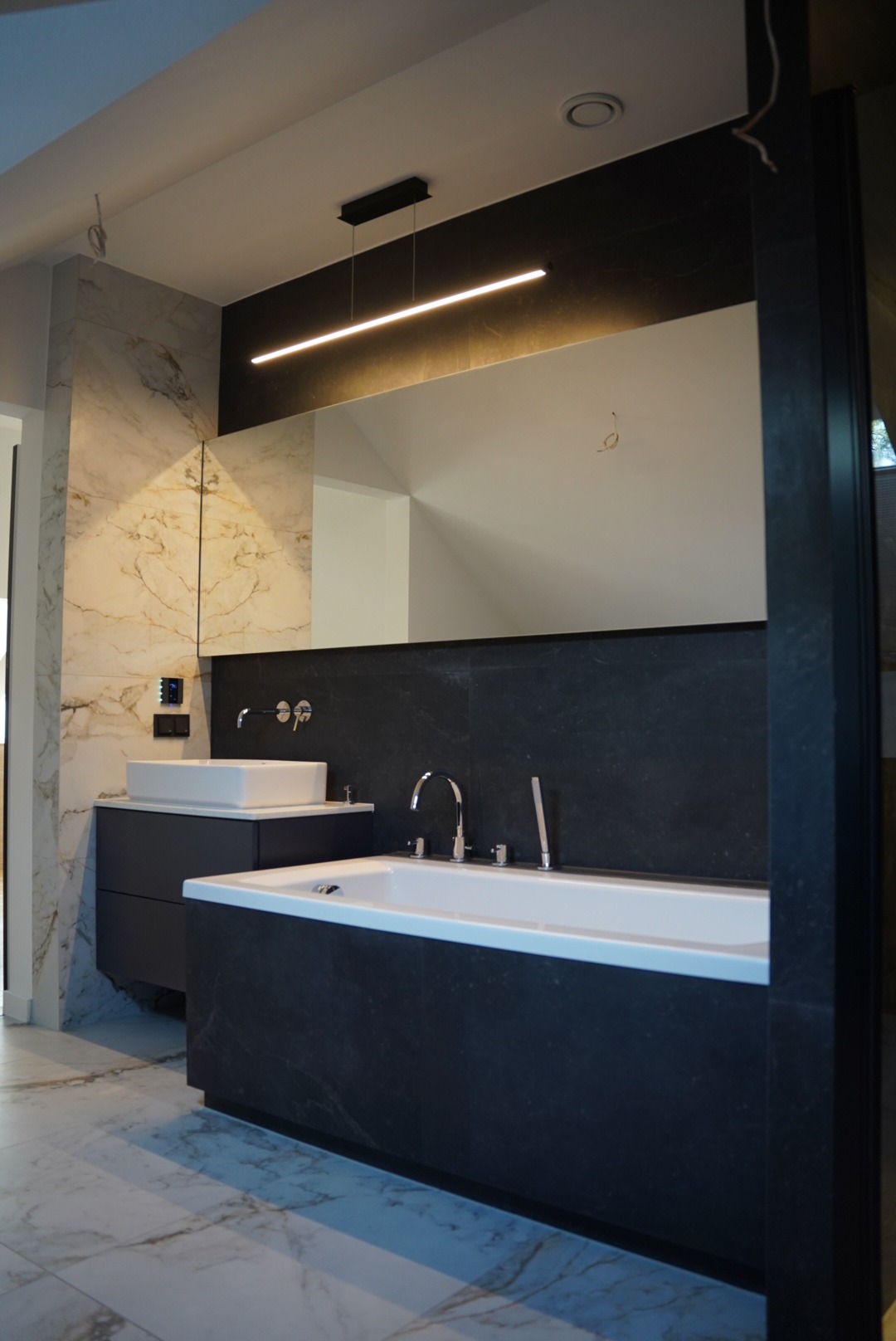Nowoczesna łazienka z marmurowymi płytkami na ścianie i podłodze, dużą wanną obudowaną czarnymi płytkami, umywalką nablatową na czarnej szafce i podłużnym oświetleniem LED nad lustrem.