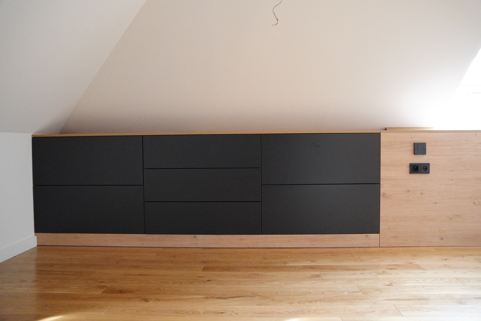Czarne, matowe fronty szafy wnękowej pod skosem, z drewnianymi elementami wykończeniowymi i gniazdkami elektrycznymi po prawej stronie, na tle drewnianej podłogi.