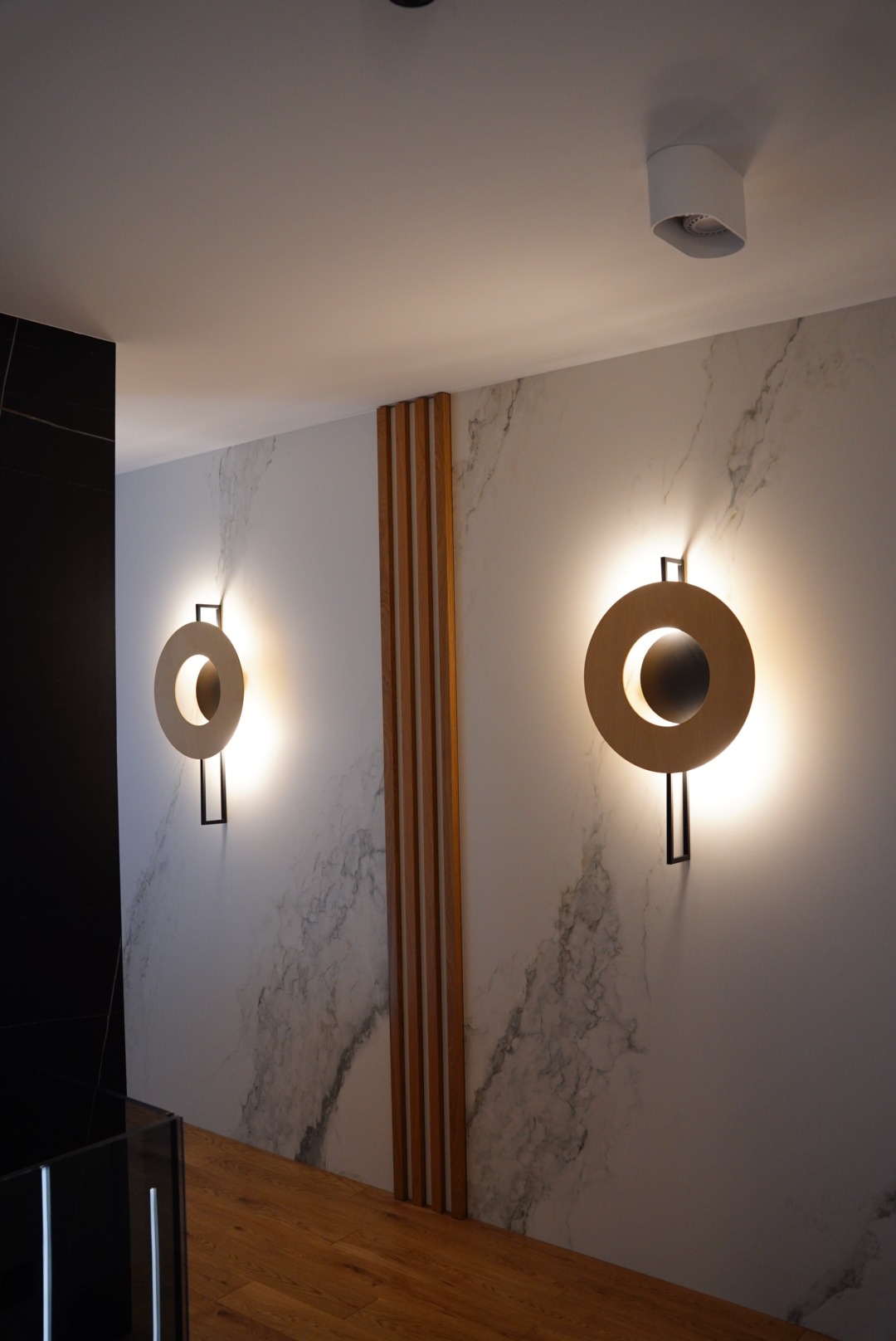 Dwa okrągłe, drewniane kinkiety z czarnymi elementami mocującymi na ścianie imitującej marmur, oddzielone pionowymi drewnianymi listwami dekoracyjnymi.
