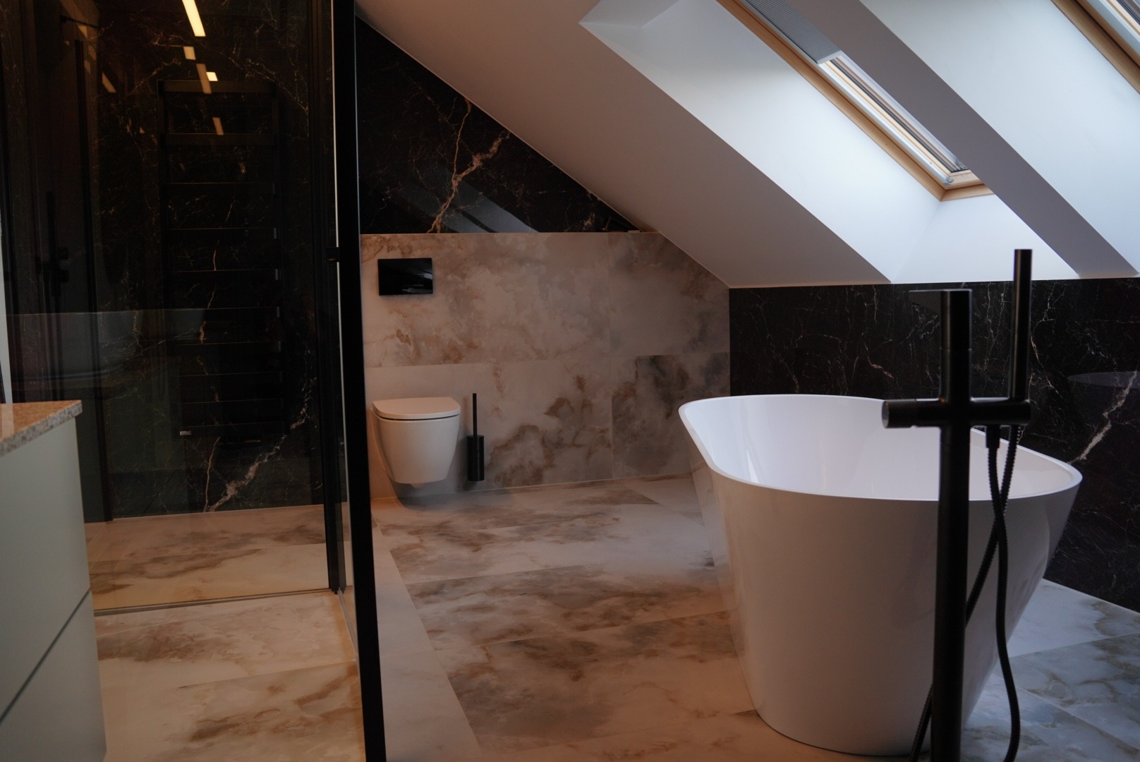 Nowoczesna łazienka na poddaszu z marmurowymi płytkami, wolnostojącą białą wanną i czarną armaturą, oświetlona światłem wpadającym przez okno dachowe.