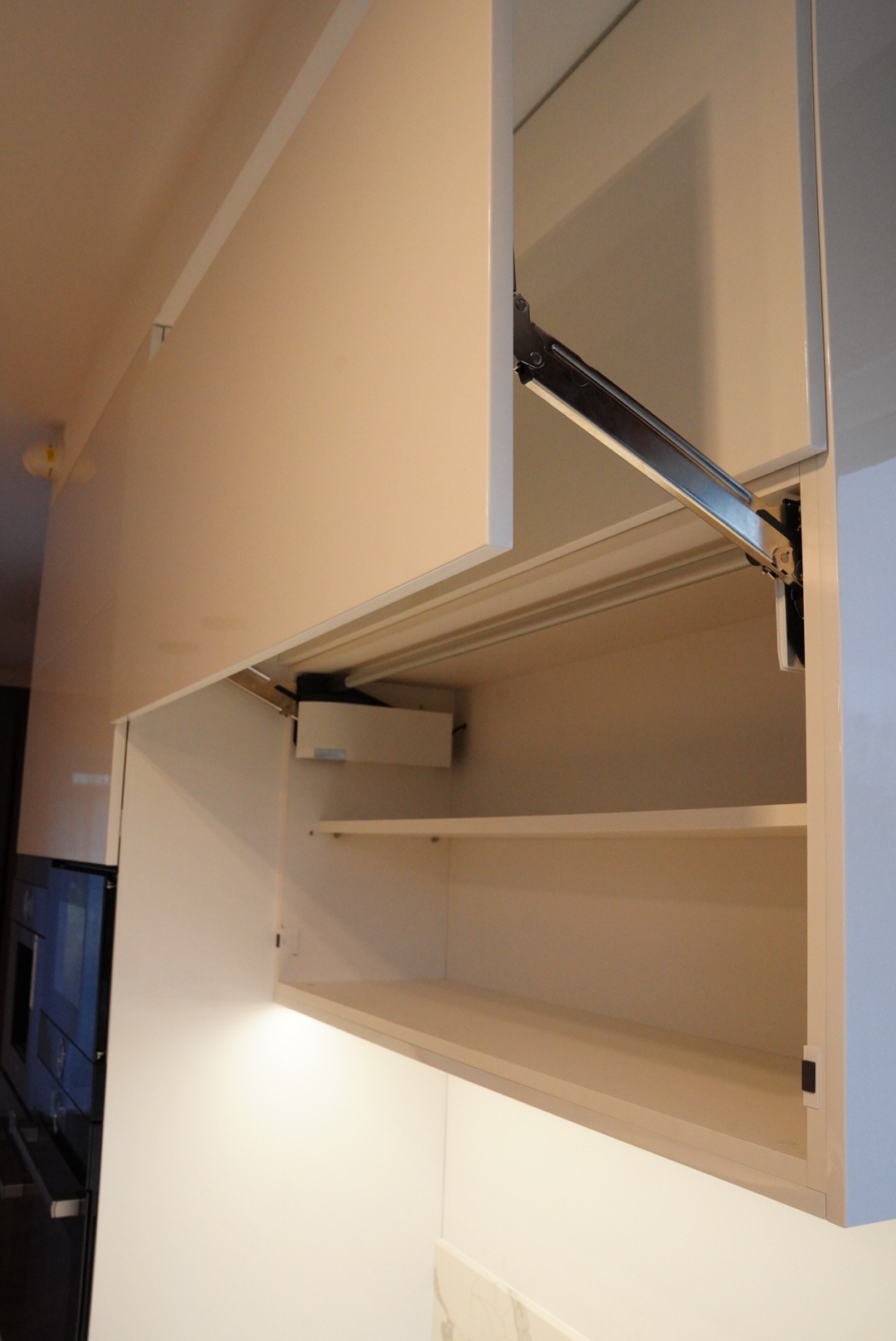 Biała szafka kuchenna wisząca z otwartymi drzwiczkami, widoczne półki wewnętrzne i mechanizm podnoszenia frontu.