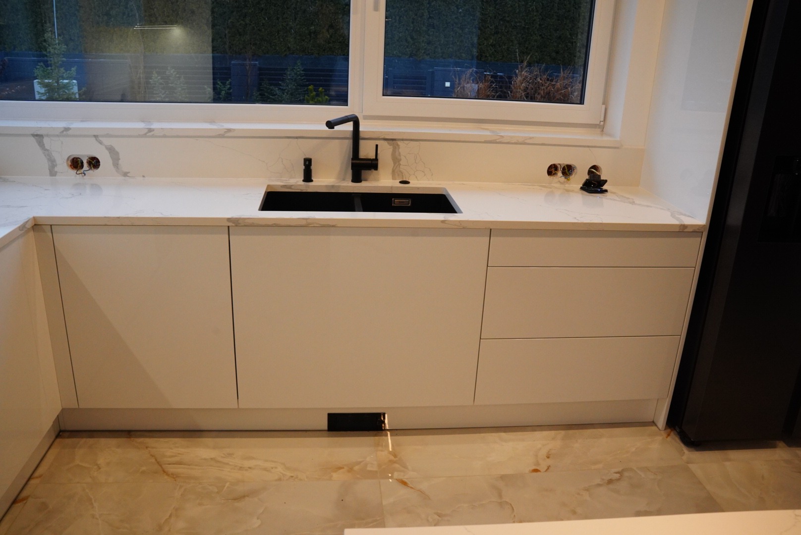 Nowoczesna kuchnia: białe szafki z blatem imitującym marmur, czarny zlew i bateria, płytki podłogowe w odcieniach beżu i brązu, okno z widokiem na zieleń.