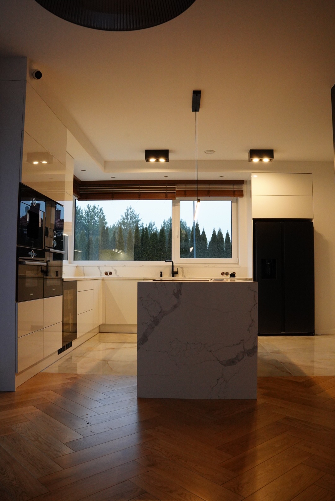 Nowoczesna kuchnia z wyspą wykończoną marmurem, drewnianą podłogą w jodełkę i dużym oknem z widokiem na zielone drzewa.