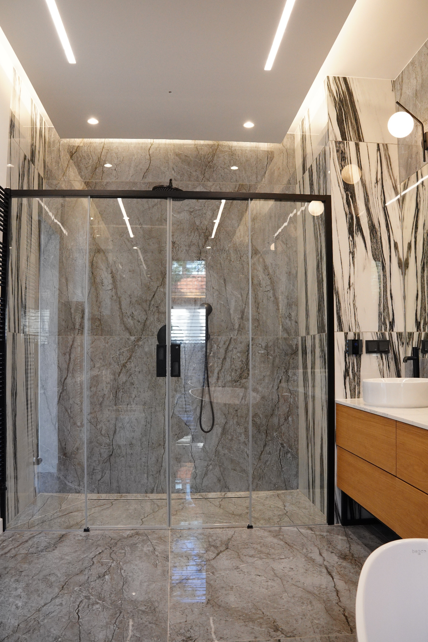 Nowoczesna łazienka z kabiną prysznicową typu walk-in, wykończona płytkami imitującymi marmur w odcieniach szarości i bieli, z czarną armaturą i drewnianą szafką pod umywalką.