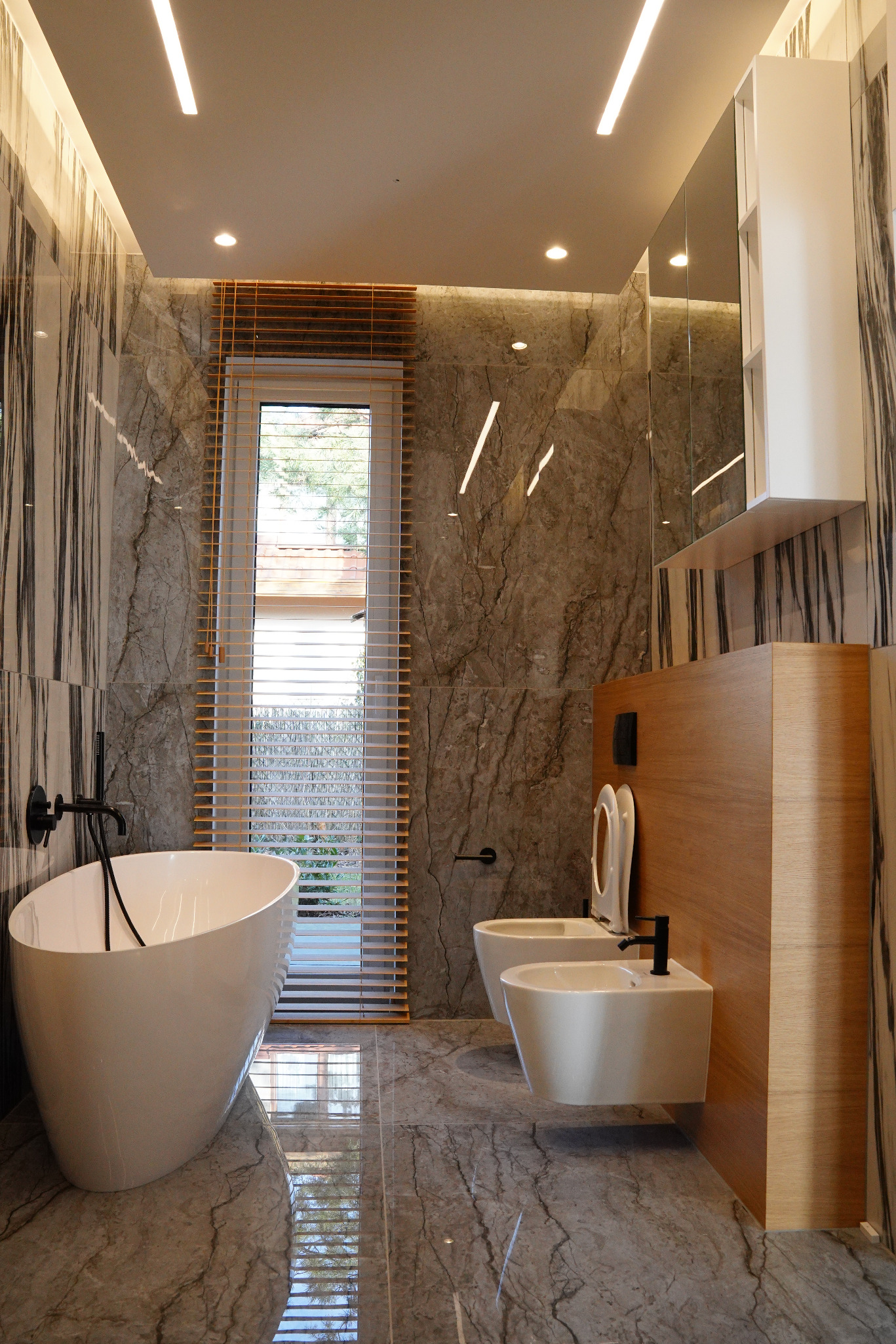Nowoczesna łazienka z marmurowymi płytkami, wolnostojącą wanną, podwieszaną toaletą i bidetem, oświetleniem LED i drewnianymi żaluzjami w oknie.