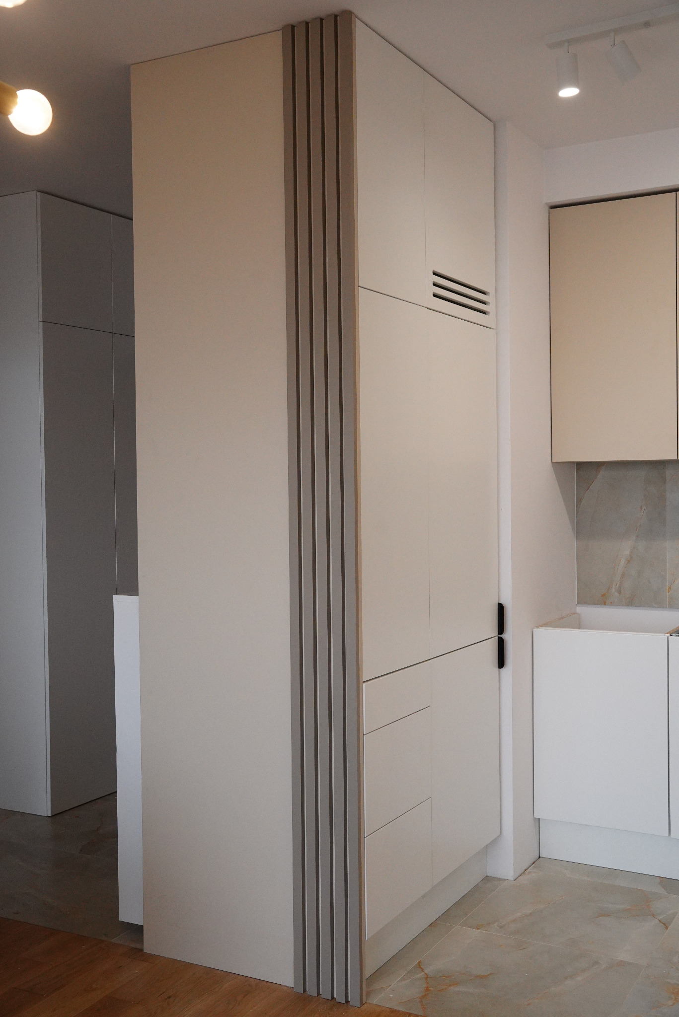 Nowoczesna, kremowa zabudowa kuchenna z pionowymi, szarymi listwami dekoracyjnymi, widoczny fragment blatu i marmurowe płytki na ścianie.