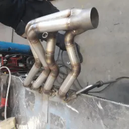 Spawany kolektor wydechowy ze stali nierdzewnej, widoczne spoiny na rurach, leżący na metalowym stole warsztatowym, w tle sprzęt spawalniczy.