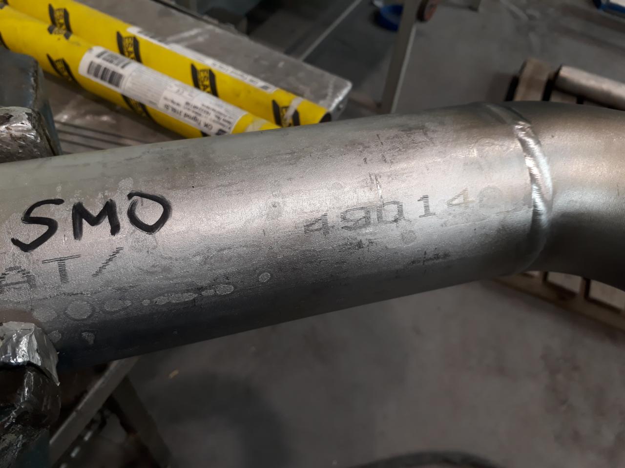 Metalowa rura ze śladami spawania, oznaczona napisem 'SMO AT/' i numerem '490140', leżąca na warsztatowym stole.