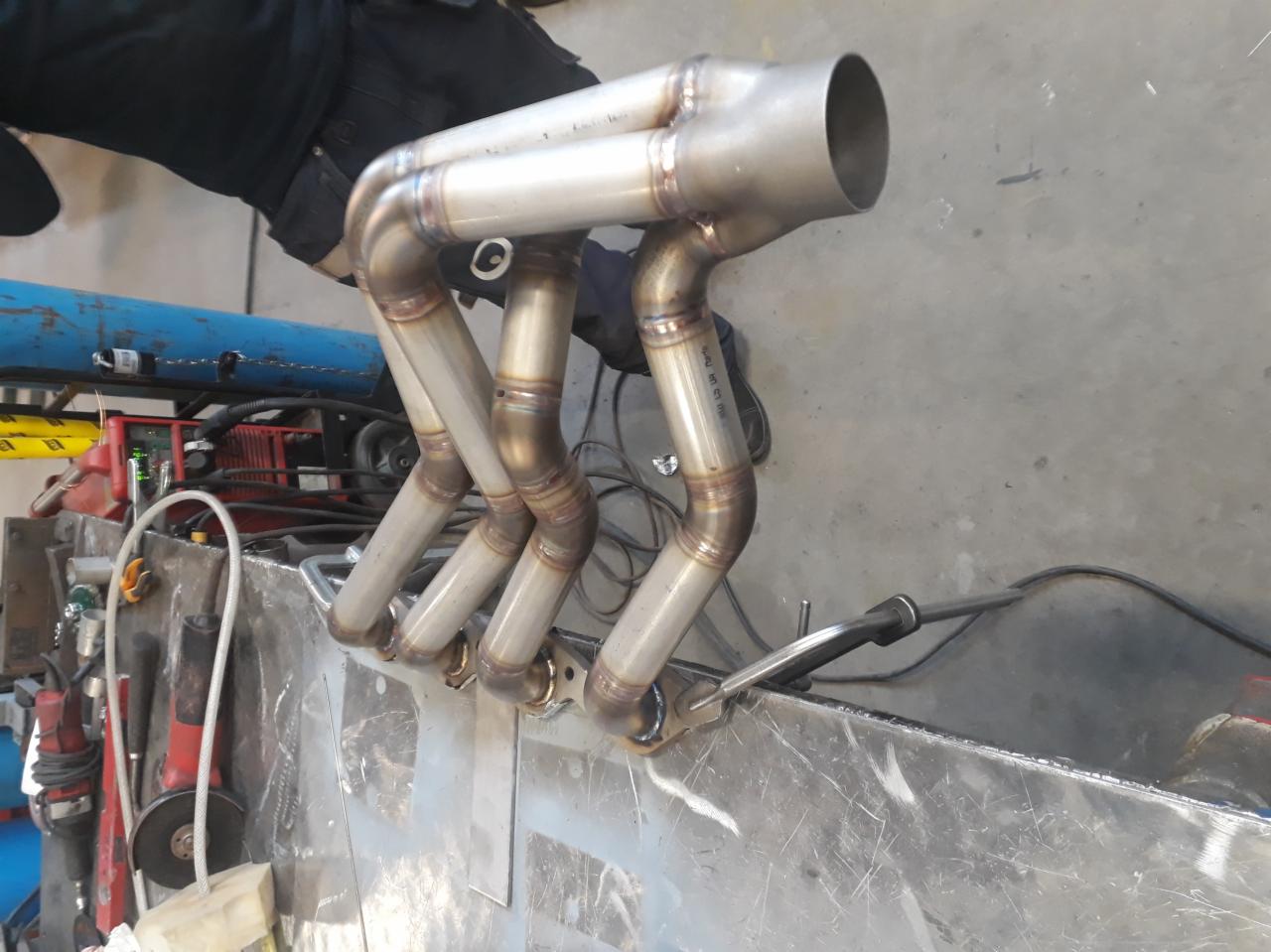 Spawany kolektor wydechowy ze stali nierdzewnej, widoczne spoiny na rurach, leżący na metalowym stole warsztatowym, w tle sprzęt spawalniczy.