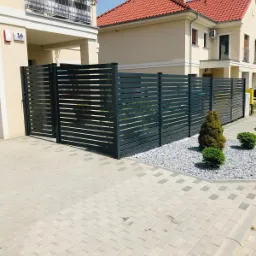 Nowoczesne, poziome ogrodzenie panelowe w kolorze grafitowym w Świebodzinie, z bramą wjazdową, otoczone białym żwirem i niskimi krzewami iglastymi, na tle jasnej zabudowy bliźniaczej z czerwonym...