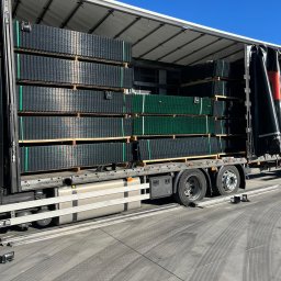 PPHU OBIS sc Jerzy Obara, Jakub Obara - Ciężarówka z otwartą naczepą załadowana paletami z panelami ogrodzeniowymi w kolorze grafitowym i zielonym, zabezpieczonymi zielonymi pasami.