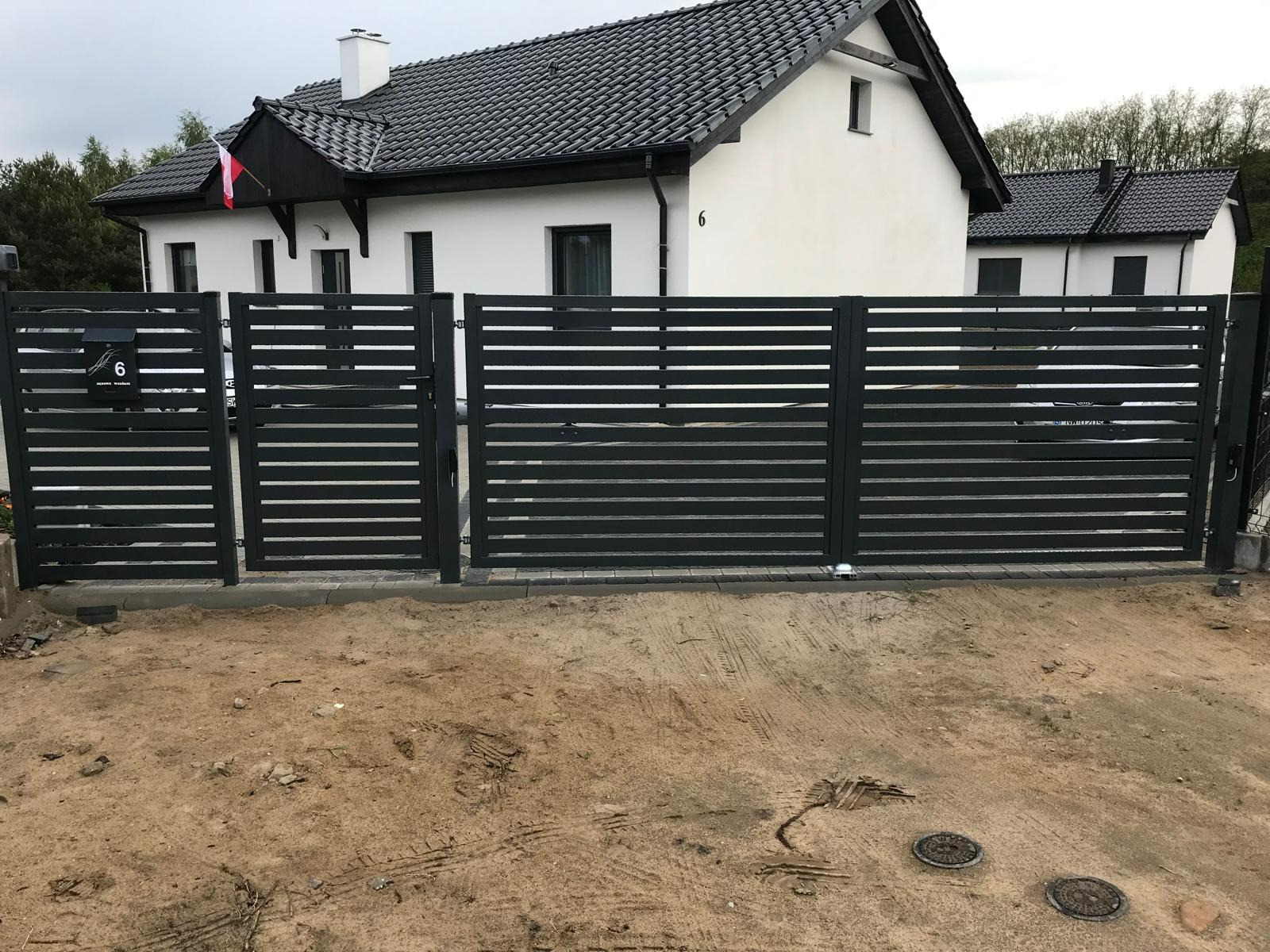 Nowoczesna, pozioma, czarna brama wjazdowa i furtka z numerem domu '6' na tle białego domu z ciemnym dachem i polską flagą.