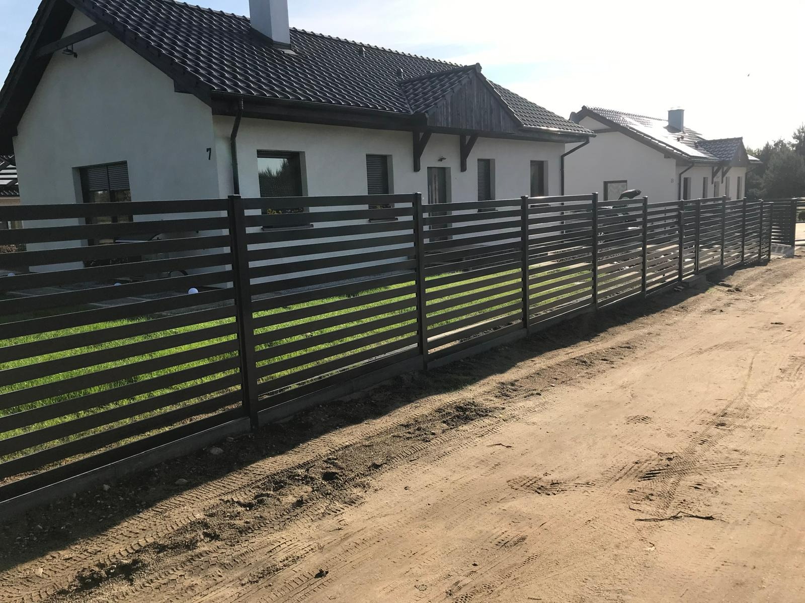 Nowoczesne poziome przęsła z metalu w kolorze grafitowym, tworzące ogrodzenie posesji z domem jednorodzinnym w tle, widoczne ślady opon na piaszczystej drodze.