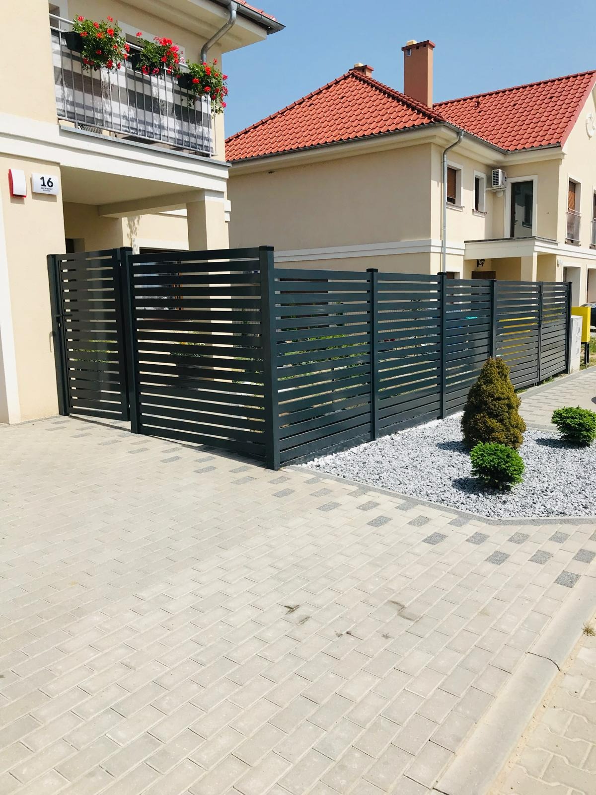 Nowoczesne, poziome ogrodzenie panelowe w kolorze grafitowym w Świebodzinie, z bramą wjazdową, otoczone białym żwirem i niskimi krzewami iglastymi, na tle jasnej zabudowy bliźniaczej z czerwonym...