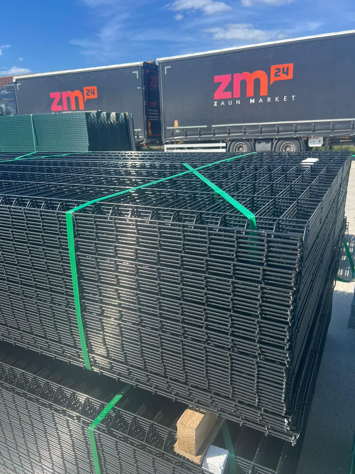 Stos czarnych paneli ogrodzeniowych związanych zielonymi taśmami transportowymi, na tle zaparkowanej ciężarówki z logo firmy.