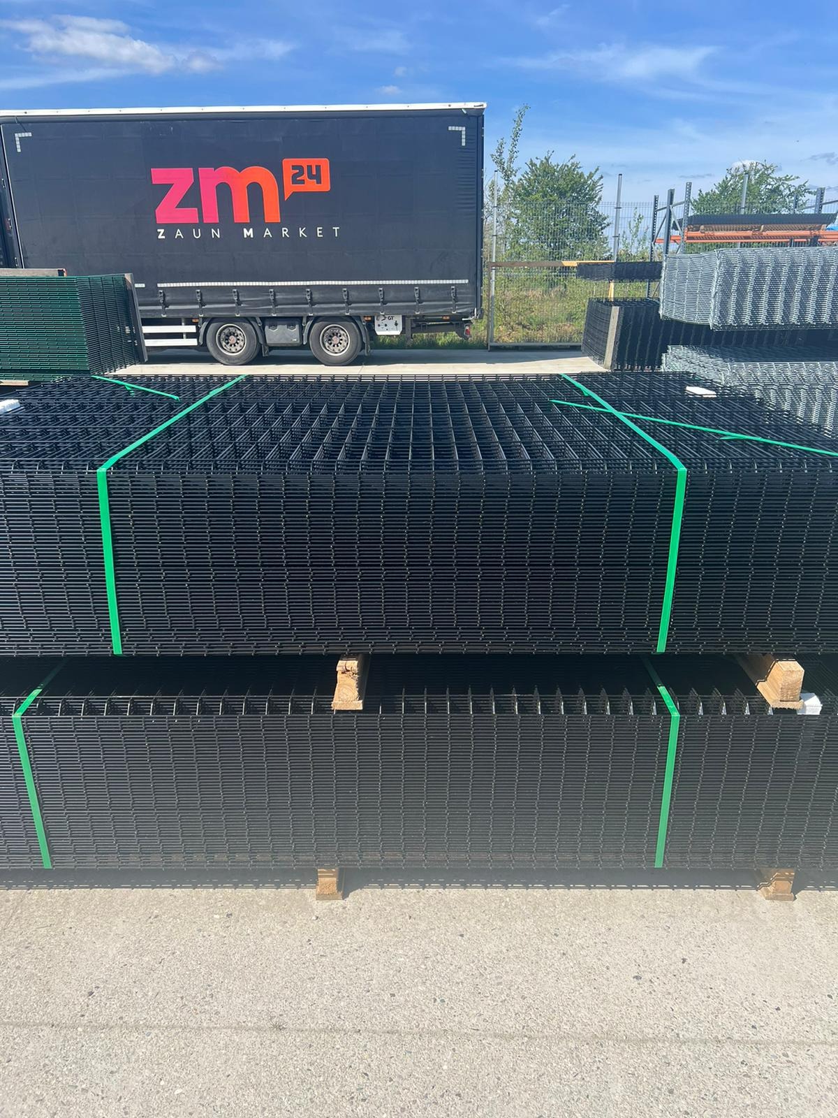 Stosy czarnych paneli ogrodzeniowych na paletach, zabezpieczonych zielonymi taśmami, z ciężarówką firmy Zaun Market w tle.