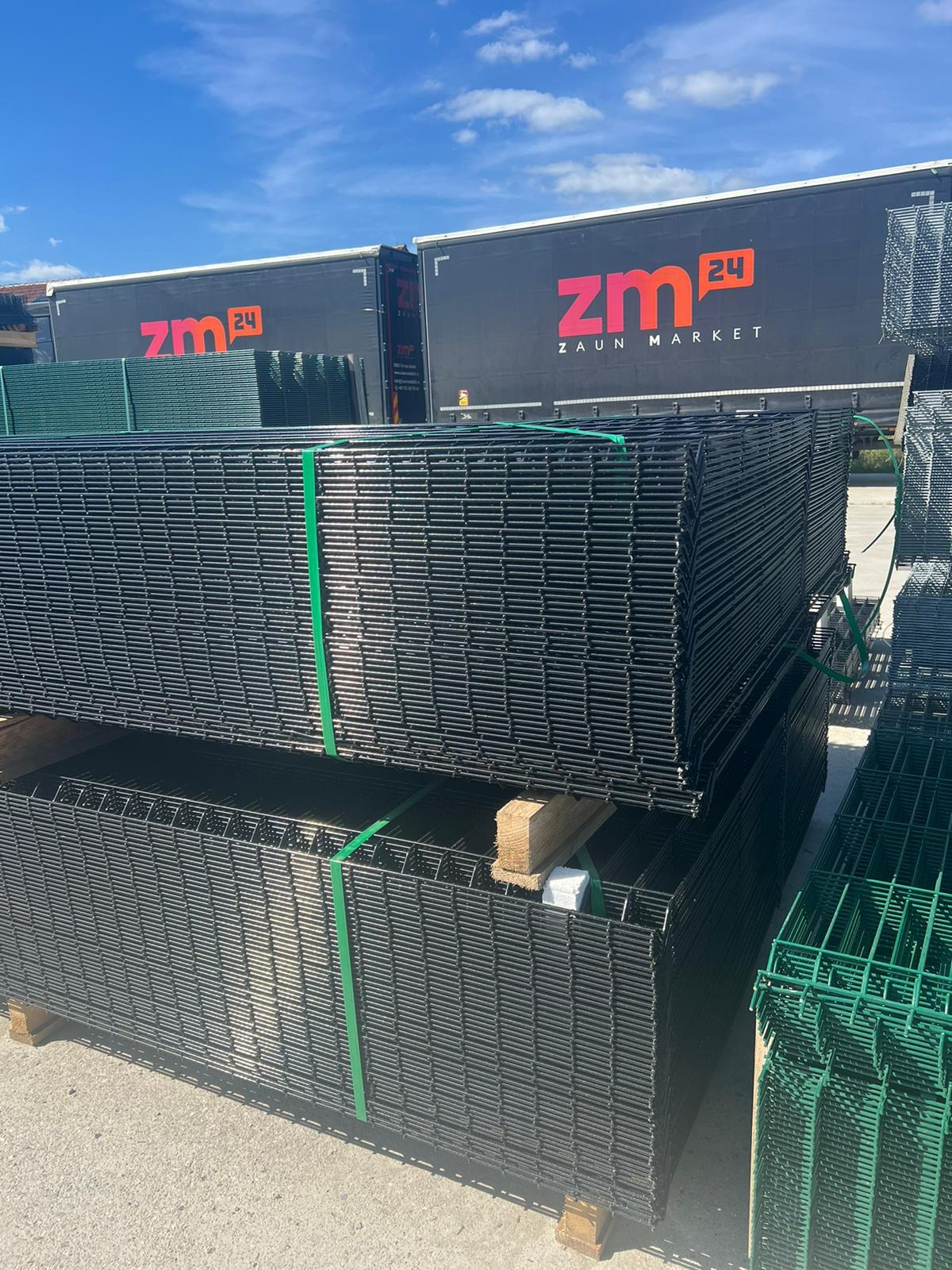 Stosy czarnych i zielonych paneli ogrodzeniowych na paletach, zabezpieczonych zielonymi taśmami, z logo firmy 'ZM 24' widocznym na naczepie ciężarówki w tle.