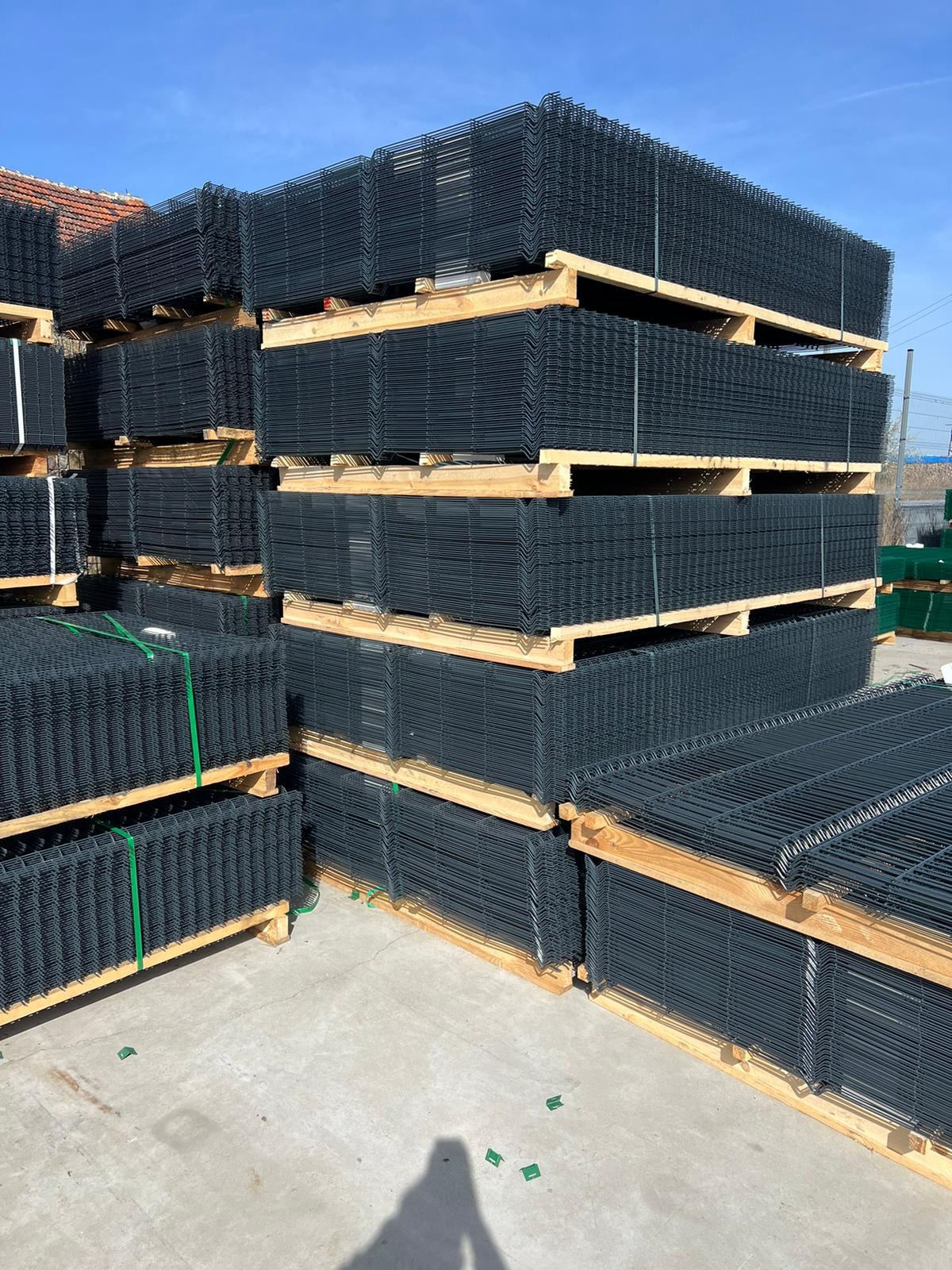 Stosy paneli ogrodzeniowych z drutu zgrzewanego, ułożone na drewnianych paletach, zabezpieczone zieloną taśmą, czekają na transport.