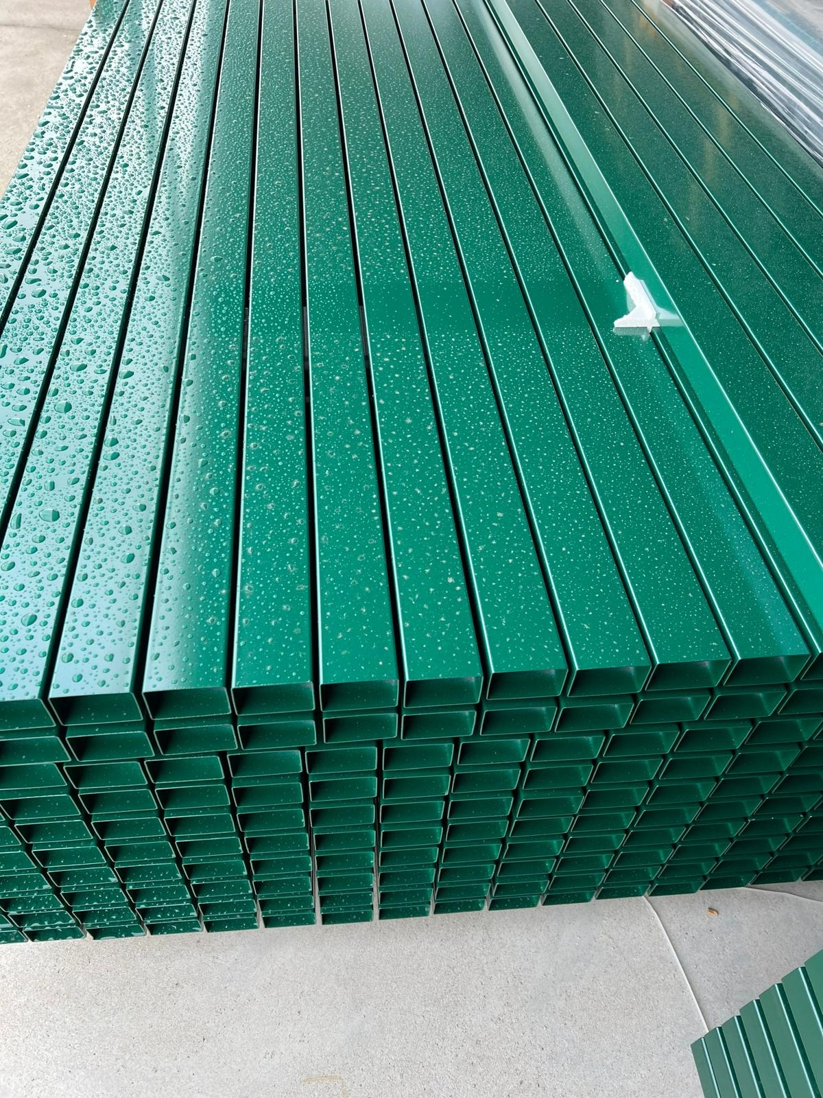 Stos zielonych, metalowych profili zamkniętych, pokrytych kroplami wody, ułożonych równolegle na betonowej powierzchni.