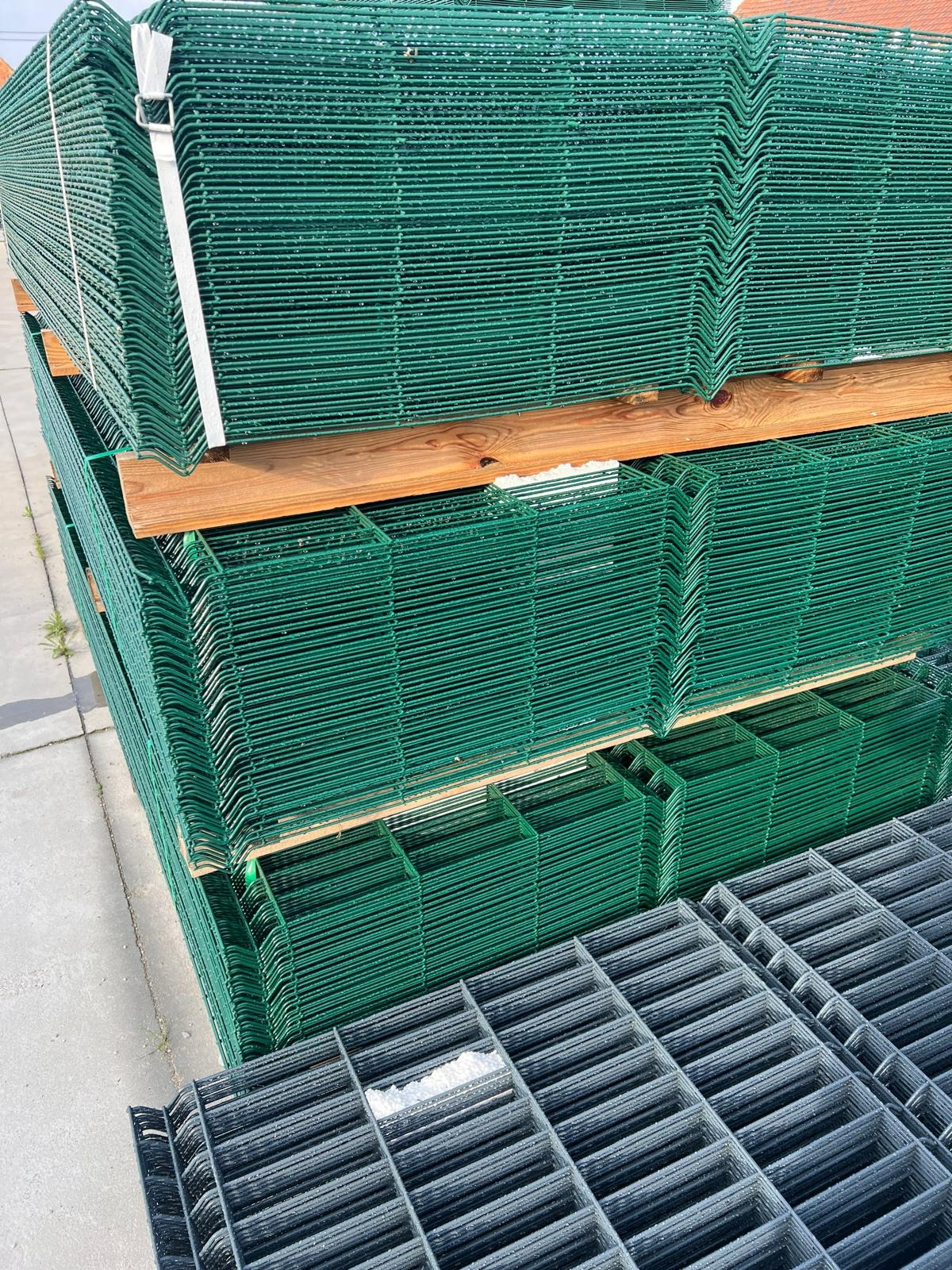 Stos paneli ogrodzeniowych w kolorze zielonym, na drewnianych belkach, obok ułożone panele siatki ogrodzeniowej w kolorze szarym.