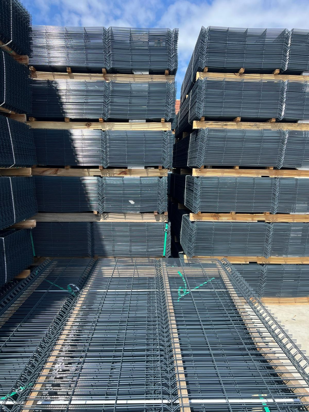 Stosy paneli ogrodzeniowych zgrzewanych z drutu, ułożone na drewnianych paletach, zabezpieczone zieloną taśmą, widoczne niebieskie niebo.