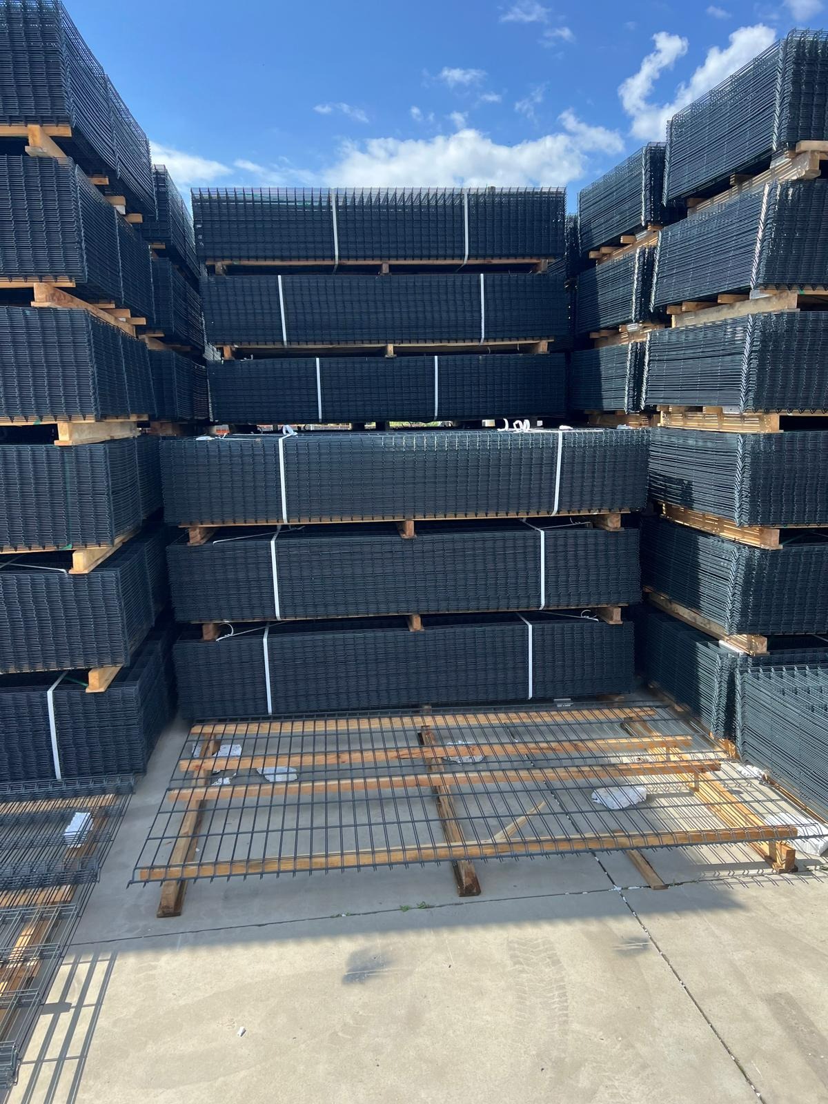 Stosy czarnych paneli ogrodzeniowych zabezpieczonych białymi taśmami, ułożone na drewnianych paletach pod błękitnym niebem z chmurami.