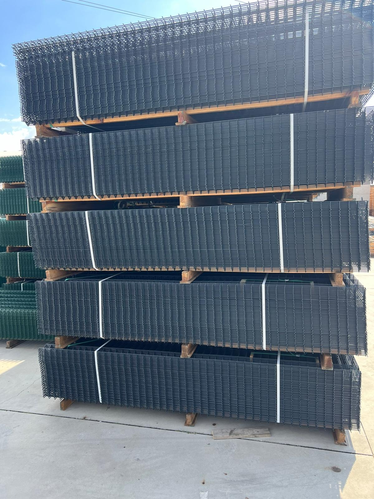 Stosy czarnych paneli ogrodzeniowych na drewnianych paletach, zabezpieczone białymi taśmami, składowane na betonowym placu.